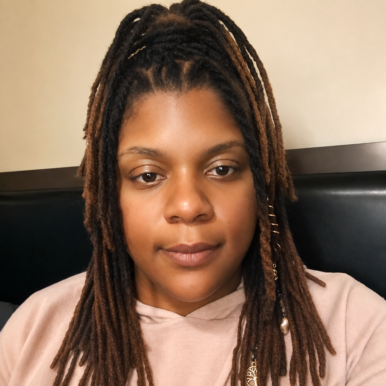 Tabitha at home, wearing a dusty pink hoodie, with long locs styled up and accented with gold jewelry